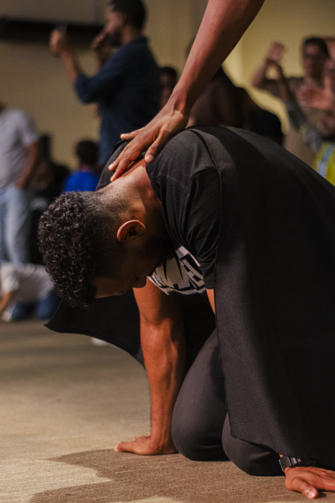Participante ajoelhado, com as mãos apoiadas no chão, recebe apoio de outra pessoa.