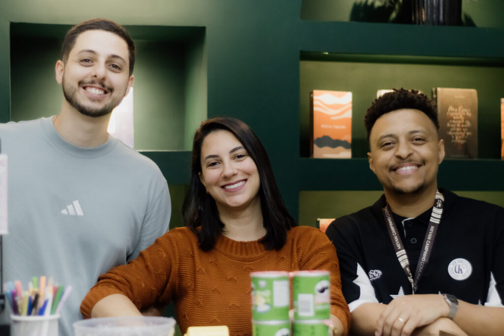 Três participantes sorrindo atrás de uma mesa de recepção.
