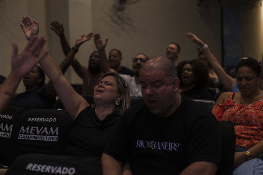 Participantes durante um culto, com uma mulher de olhos fechados e o braço levantado em primeiro plano.
